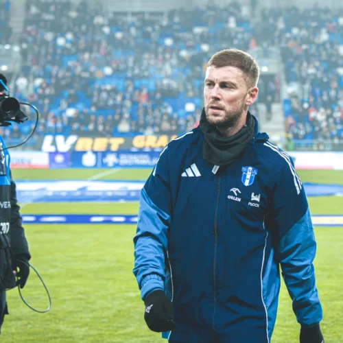 Na zdjęciu znajduje się mężczyzna w odzieży sportowej, idący po boisku piłkarskim, podczas gdy w tle widać tłum kibiców oraz kamerzystę rejestrującego wydarzenie.