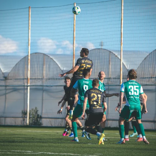 Na zdjęciu widoczna jest scena z meczu piłkarskiego, gdzie zawodnik w czarnej koszulce skacze, próbując przejąć piłkę nad grupą graczy w zielonych strojach.