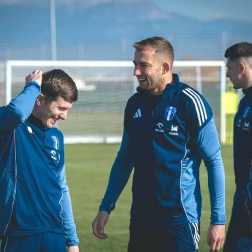 Na zdjęciu widać grupę piłkarzy w niebieskich strojach treningowych, którzy rozmawiają i śmieją się na boisku treningowym.