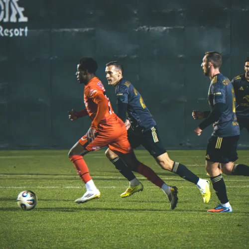 Na boisku piłkarskim dwóch zawodników w pomarańczowych strojach rywalizuje z grupą graczy w granatowych koszulkach podczas intensywnej akcji meczu.