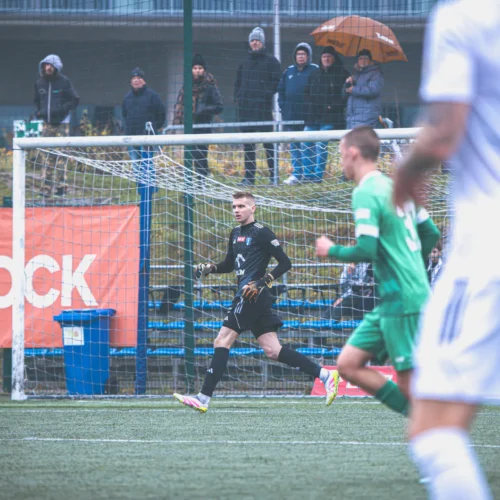 Na zdjęciu znajduje się sytuacja z meczu piłkarskiego, w której bramkarz w czarnym stroju biegnie w kierunku bramki, podczas gdy zawodnicy w zielonym i białym stroju są w ruchu, a w tle widać widownię.