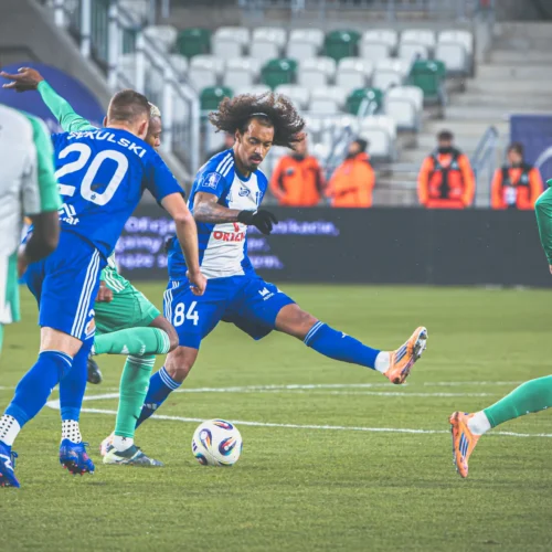 Na zdjęciu dwóch zawodników w niebieskich strojach stara się przejąć piłkę, podczas gdy gracz w zielonym biega w stronę bramki, a na trybunach w tle widać kibiców.