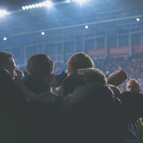 Grupa ludzi cieszy się w emocjonalnym uścisku podczas meczu piłkarskiego na stadionie.