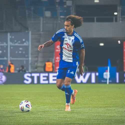 Na zdjęciu widać piłkarza w niebieskiej koszulce, który biega z piłką na boisku podczas meczu.