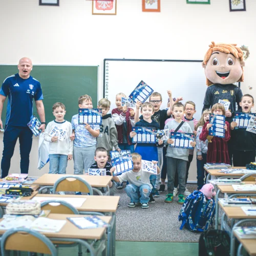 Na zdjęciu w klasie szkolnej dzieci trzymają nagrody i pozują z postacią maskotki oraz nauczycielem.