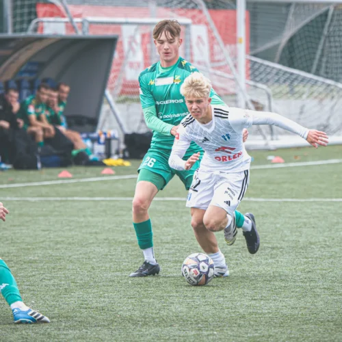 Na zdjęciu widać dwóch młodych piłkarzy rywalizujących o piłkę na boisku, z jednym zawodnikiem w białym stroju mknącym w kierunku bramki, podczas gdy drugi zawodnik w zielonym stroju próbuje go powstrzymać.