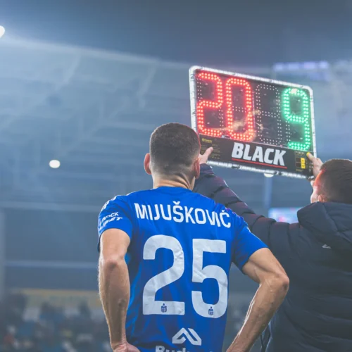 Na zdjęciu gracz w niebieskiej koszulce z numerem 25 stoi na boisku, podczas gdy sędzia trzyma tablicę z numerami 20 i 9, wskazując na zmianę zawodnika.