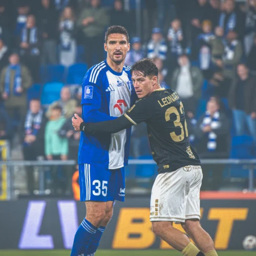 Zawodnicy w strojach piłkarskich stoją blisko siebie na boisku, a w tle widać widownię.