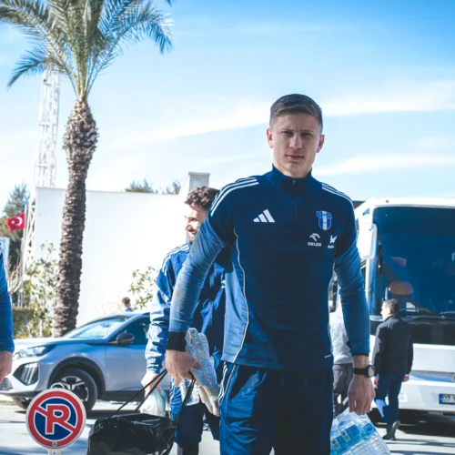 Na zdjęciu widać trzech mężczyzn w niebieskich strojach sportowych, którzy idą w kierunku autobusu na tle palm i błękitnego nieba.