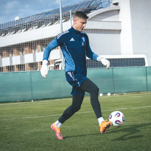 Na zdjęciu widoczny jest bramkarz w niebieskim stroju treningowym, który technicznie kopie piłkę na zielonym boisku.