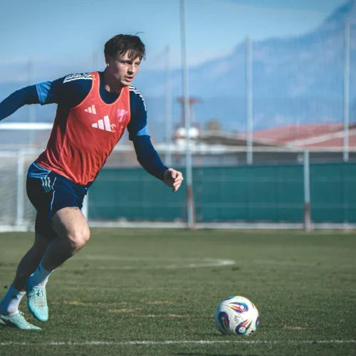 Na zdjęciu widoczny jest młody mężczyzna w czerwonej koszulce treningowej, biegnący po boisku piłkarskim w kierunku piłki.