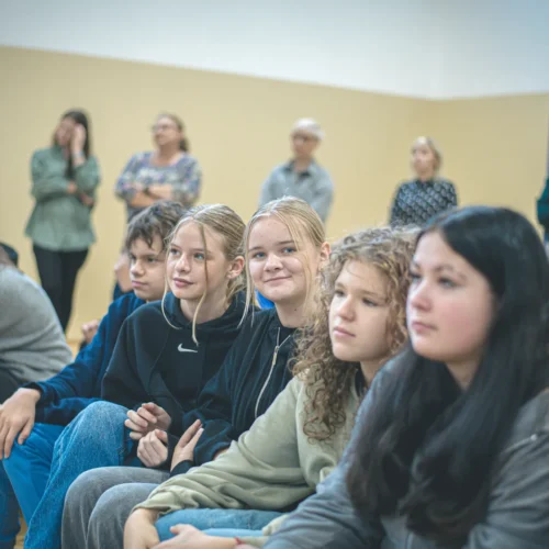 Grupa młodych ludzi siedzi w rządku, skupiona na czymś, z różnymi wyrazami twarzy.
