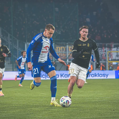 Na zdjęciu widać dynamiczną akcję na boisku piłkarskim z zawodnikami w niebieskich i czarnych strojach, którzy walczą o piłkę.