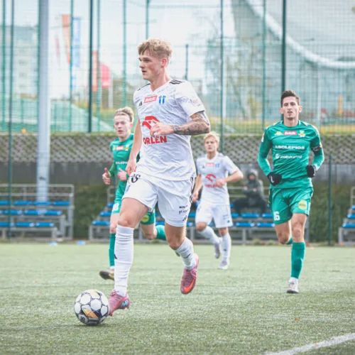 Mężczyzna w białej koszulce piłkarskiej biegnie z piłką na boisku, podczas gdy dwóch gnatych zawodników w zielonych strojach podąża za nim.