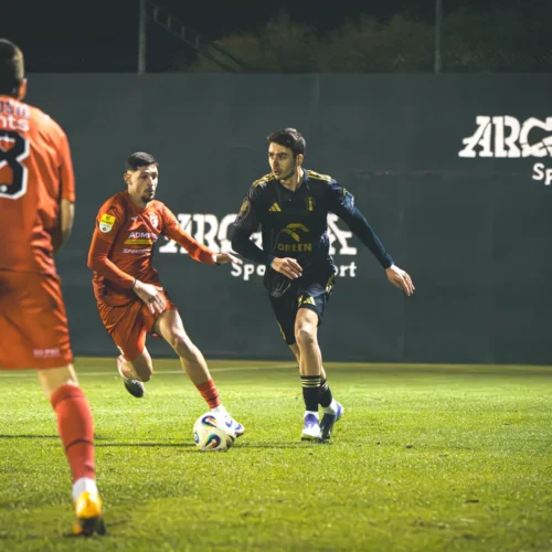 Na zdjęciu przedstawieni są dwaj piłkarze grający w trakcie meczu na boisku, jeden z nich w czerwonym stroju, a drugi w czarnym, z piłką na zielonej murawie.