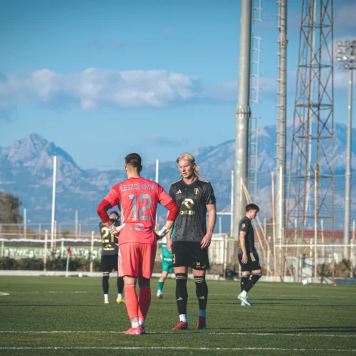 Na zdjęciu znajduje się dwóch piłkarzy na boisku, z tłem górskim oraz innymi zawodnikami w oddali.
