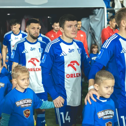 Na zdjęciu widać piłkarzy wychodzących na boisko, trzymających za ręce dzieci w niebieskich koszulkach, w tle widoczna jest publika oraz inne postacie.