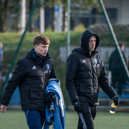 Dwaj mężczyźni w odzieży sportowej idą po boisku piłkarskim, jeden z nich trzyma butelkę napoju.