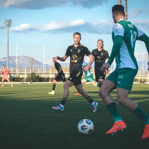 Na zdjęciu widać zawodników grających w piłkę nożną na boisku, z górami w tle i słoneczną pogodą.