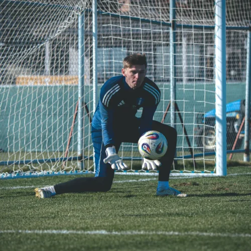 Na zdjęciu widać bramkarza w niebieskim stroju, który przygotowuje się do złapania piłki w trakcie treningu na boisku piłkarskim.