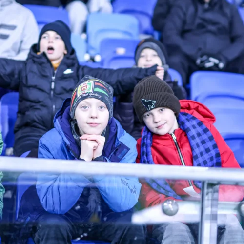 Na zdjęciu widać grupę dzieci siedzących na trybunach, z wyraźnie widocznymi emocjami podczas wydarzenia sportowego.