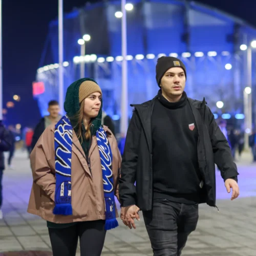 Para spaceruje trzymając się za ręce w nocy, w otoczeniu oświetlonych budynków i przechodniów.