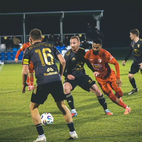 Na zdjęciu widać piłkarzy w trakcie meczu na zielonym boisku, gdzie zawodnicy w czarnych strojach starają się odebrać piłkę graczowi w czerwonym stroju.