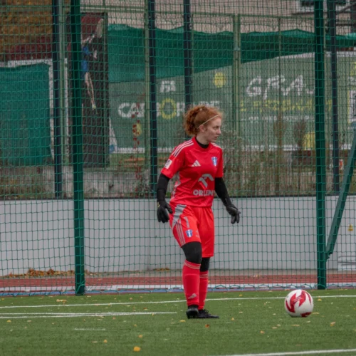 Na zdjęciu widać młodą zawodniczkę piłki nożnej w czerwonym stroju, stojącą na boisku, przygotowującą się do rozpoczęcia akcji.