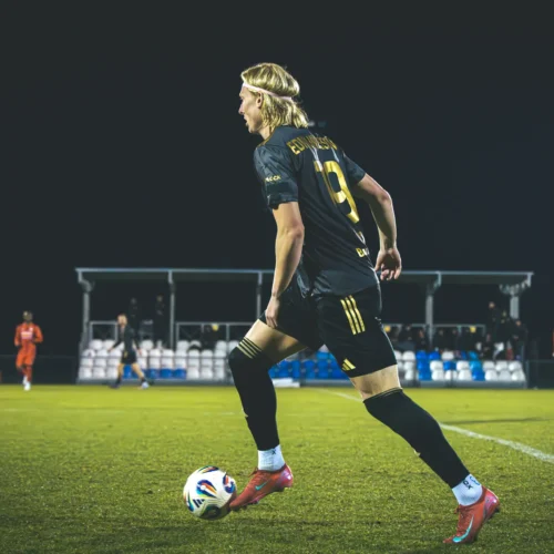 Na zdjęciu widoczny jest piłkarz w czarnym stroju, który wykonuje drybling z piłką na murawie, podczas gdy w tle odbywa się mecz piłkarski.