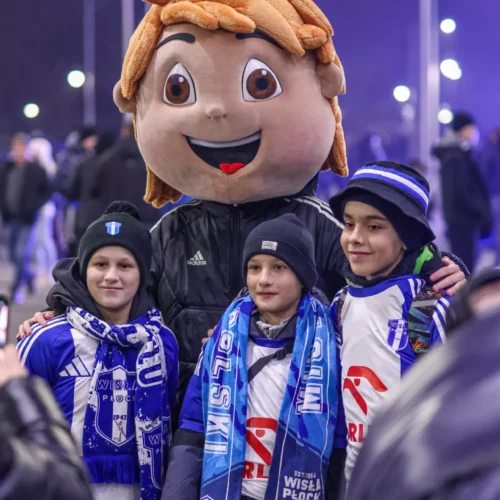Na zdjęciu widoczni są trzej chłopcy przebrani w kibicowskie koszulki i szaliki, pozujący z postacią maskotki z uśmiechem.