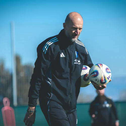 Trener piłkarski na boisku, niosący piłki w rękach, w tle jest niebieskie niebo.