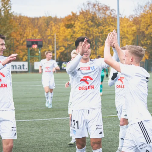 Na zdjęciu widać grupę piłkarzy w białych strojach sportowych, którzy celebrują wspólnie zdobyty gol na boisku otoczonym drzewami w kolorowych, jesiennych liściach.