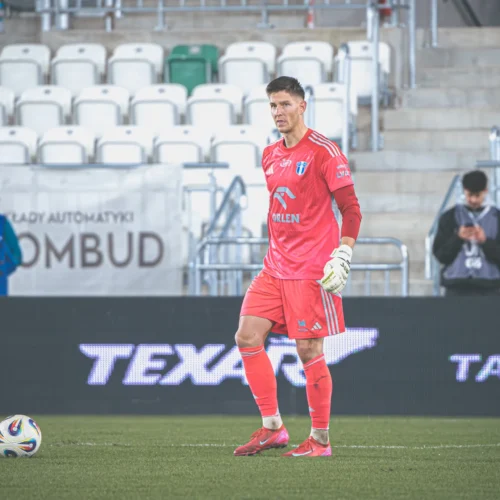 Na zdjęciu widać bramkarza w różowym stroju na boisku piłkarskim, z piłką przed sobą oraz widownią w tle.