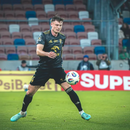 Na zdjęciu widać piłkarza w czarnej koszulce, który kontroluje piłkę podczas meczu na boisku, w tle z widownią.