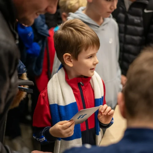 Na zdjęciu widać radosnego chłopca w czerwonej kurtce i szaliku, który trzyma kartkę i uczestniczy w grupie dzieci, z entuzjazmem oglądających coś interesującego.