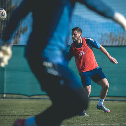 Na zdjęciu widać piłkarza w pomarańczowej koszulce treningowej, który obserwuje lecącą w jego kierunku piłkę podczas treningu na boisku.