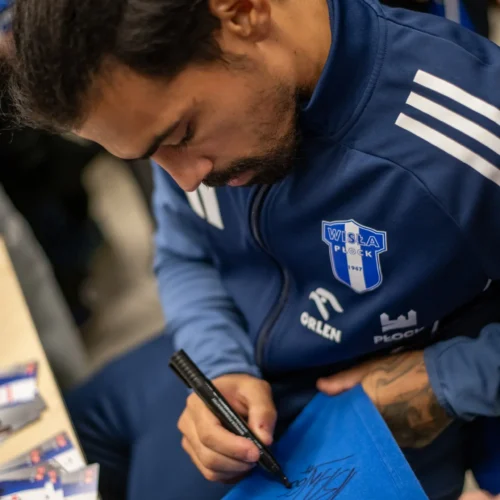 Na zdjęciu widoczny jest mężczyzna w sportowym stroju, który podpisuje niebieską chustkę.