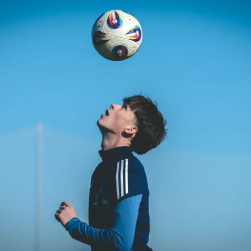 Na zdjęciu widać młodego mężczyznę, który wpatruje się w unoszącą się nad nim piłkę nożną na tle błękitnego nieba.