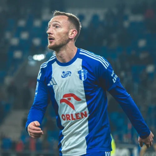 Na zdjęciu widać piłkarza w niebiesko-białej koszulce, który z zaangażowaniem uczestniczy w meczu piłkarskim.