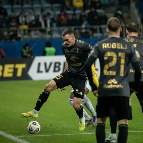 Na zdjęciu widoczna jest akcja piłkarska, w której zawodnik kopie piłkę podczas meczu, a w tle można dostrzec innych graczy oraz kibiców na trybunach.