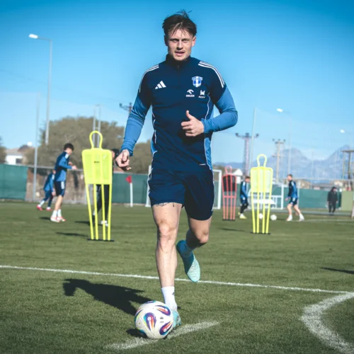 Na zdjęciu widać piłkarza biegnącego po boisku treningowym, z innymi zawodnikami w tle, w słoneczny dzień.