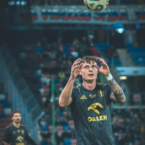 Na zdjęciu widoczny jest zawodnik w czarnej koszulce sportowej, który skupia się na piłce unoszącej się w powietrzu podczas meczu piłkarskiego.