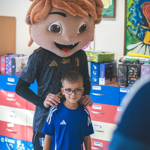 Na zdjęciu widać dziecko w niebieskiej koszulce stojące przed osobą w kostiumie maskotki z dużą głową, w kolorowej sali pełnej zabawek i kolorowych szafek.