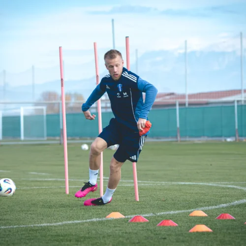 Na zdjęciu dwóch piłkarzy trenuje na boisku, wykonując ćwiczenia ze słupkami i pachołkami.
