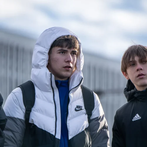 Trzech młodych mężczyzn w kurtkach zimowych rozmawia na zewnątrz, z wyrazami skupienia na twarzach.