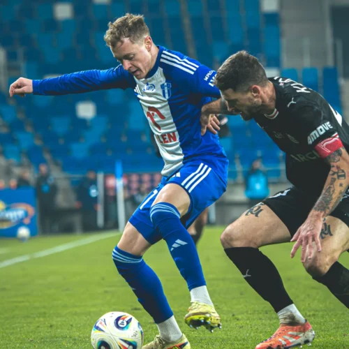 Na zdjęciu dwóch piłkarzy walczy o piłkę na boisku, jeden z nich jest ubrany w niebiesko-białą koszulkę, a drugi w czarną.