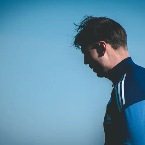 Na zdjęciu widoczny jest profil mężczyzny w niebieskiej odzieży sportowej, stojącego na tle jasnego, błękitnego nieba.