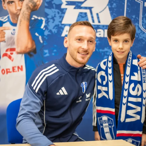 Na zdjęciu widać uśmiechniętego mężczyznę w dresie oraz chłopca trzymającego szalik, siedzących w tle z plakatami drużyny sportowej.