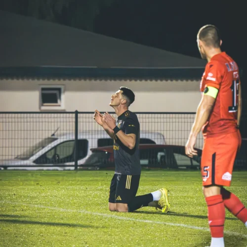 Na zdjęciu widać piłkarza klęczącego na boisku, składającego dłonie w geście modlitwy, podczas gdy drugi zawodnik w czerwonym stroju obserwuje sytuację.