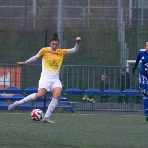 Na zdjęciu widoczne są dwie zawodniczki podczas meczu piłki nożnej, jedna w żółtej, a druga w niebieskiej koszulce.